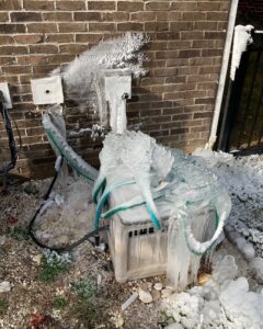 Close-up of a garden hose bib and spigot encased in thick ice against a brick wall, with a frozen garden hose coiled over an ice-covered outdoor unit, illustrating burst pipe damage from not disconnecting a hose before winter