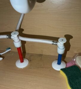 Under-sink view of color-coded red and blue push-to-connect compression valves on the hot and cold supply lines, next to a toothbrush, sponge, and soap on a wood cabinet shelf.