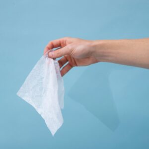 Close-up of a hand holding a single crumpled white flushable wipe against a light blue background.