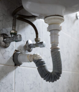 Underside of a bathroom sink showing a flexible accordion-style drain trap connecting the sink's drain pipe to the wall outlet, alongside the shutoff valves and supply lines.