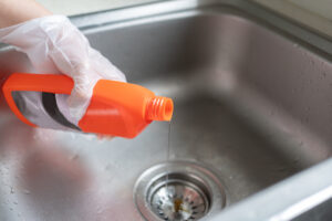 A gloved hand pours liquid chemical drain cleaner from an orange bottle into a stainless steel kitchen sink drain.